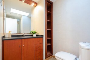 Un baño con tocador de madera, lavamanos, espejo, inodoro y estante.