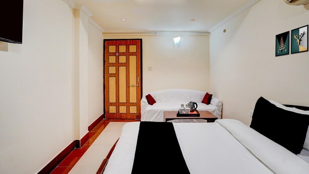 A hotel room with a bed, a sofa, a small table with a tray, a wooden door, and a wall-mounted TV. - Deluxe Banshi Bhawan (Prayagraj)
