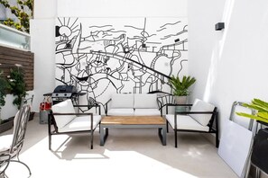 Ein moderner Außenbereich zum Sitzen mit minimalistischem Design, der eine Sitzecke mit einer Couch und Sesseln, einen Holztisch sowie eine große, abstrakte Wandmalerei umfasst.