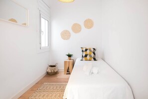 A minimalist bedroom with a bed, a small bedside table, a plant, and a window. - Cute Apartment in the Heart of Vilanova | Virgi By Palmera (Vilanova i la Geltrú)