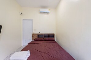 Quarto com cama, porta branca, ar-condicionado na parede e uma pequena prateleira com uma planta.