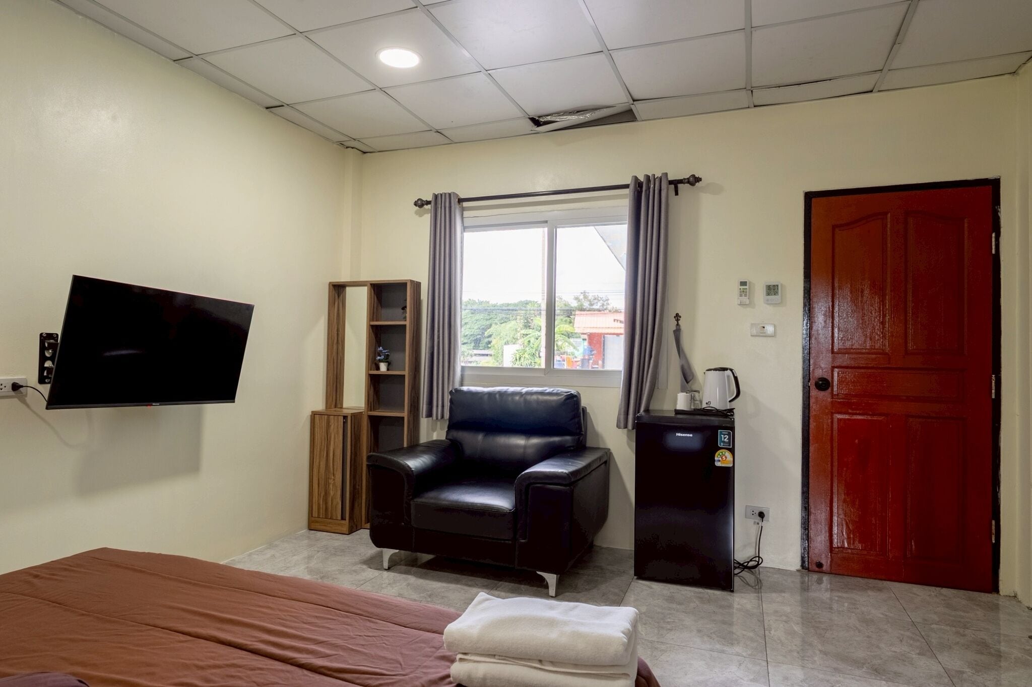 Una habitación con una cama, un sillón de cuero negro, un televisor montado en la pared, un ventanal con cortinas, una puerta roja y un pequeño escritorio con una tetera.