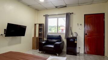 Una habitación con una cama, un sillón de cuero negro, un televisor montado en la pared, un ventanal con cortinas, una puerta roja y un pequeño escritorio con una tetera.