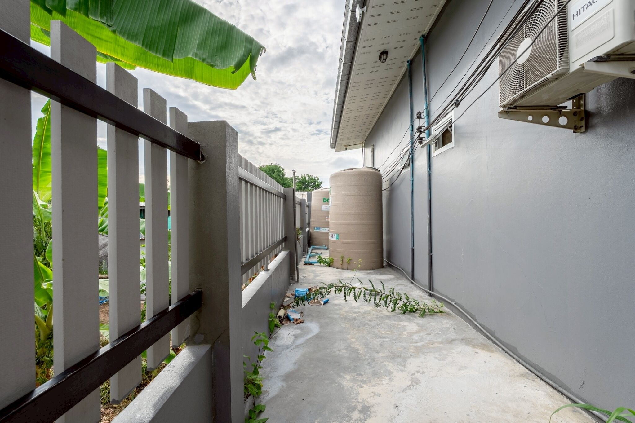 A narrow alleyway with a white fence, a large cylindrical tank, and a wall-mounted air conditioning unit.