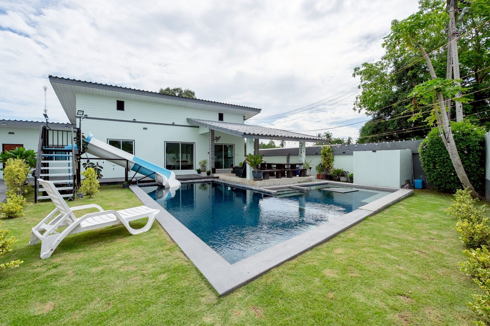 A residential swimming pool with a slide, surrounded by a well-maintained lawn and a house with a flat roof.