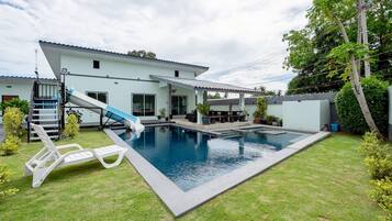 A residential swimming pool with a slide, surrounded by a well-maintained lawn and a house with a flat roof.