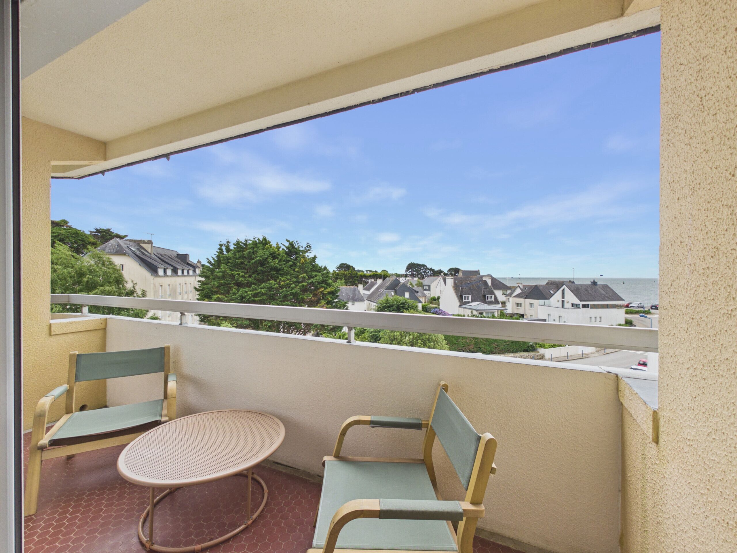 A balcony with two chairs and a small table, overlooking a residential area and the sea.