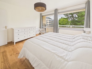 Ein Schlafzimmer mit einem großen Bett, einer weißen Kommode, einem Stuhl und Blick auf Gebäude und Bäume durch das Fenster.