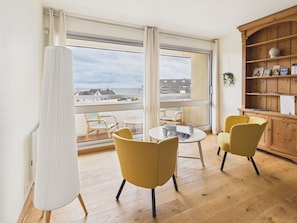 Ein Zimmer mit Balkon, einem runden Tisch, zwei gelben Stühlen, einer Holzkommode und Meerblick.