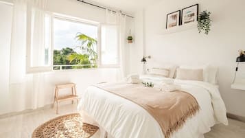 Une chambre avec une grande fenêtre, un lit recouvert de draps blancs, un petit tabouret en bois et un tapis tissé.