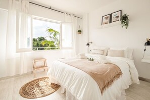 A bedroom with a large window, a bed with white bedding, a small wooden stool, and a woven rug. - Sunny & Stylish Apartment with A/C | Trini by Palmera (Sant Pere de Ribes)