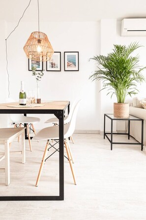 A modern dining area with a wooden table, white chairs, a hanging light fixture, framed pictures on the wall, and a potted plant. - Sunny & Stylish Apartment with A/C | Trini by Palmera (Sant Pere de Ribes)