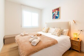 Un dormitorio con cama, almohadas, una mesita de noche con lámpara y una ventana.