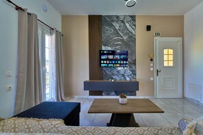 A hotel room with a bed, a TV mounted on the wall, a table with a bowl of fruit, and a door with a light above it. - Villa Escapes By The Beach (Sidari)