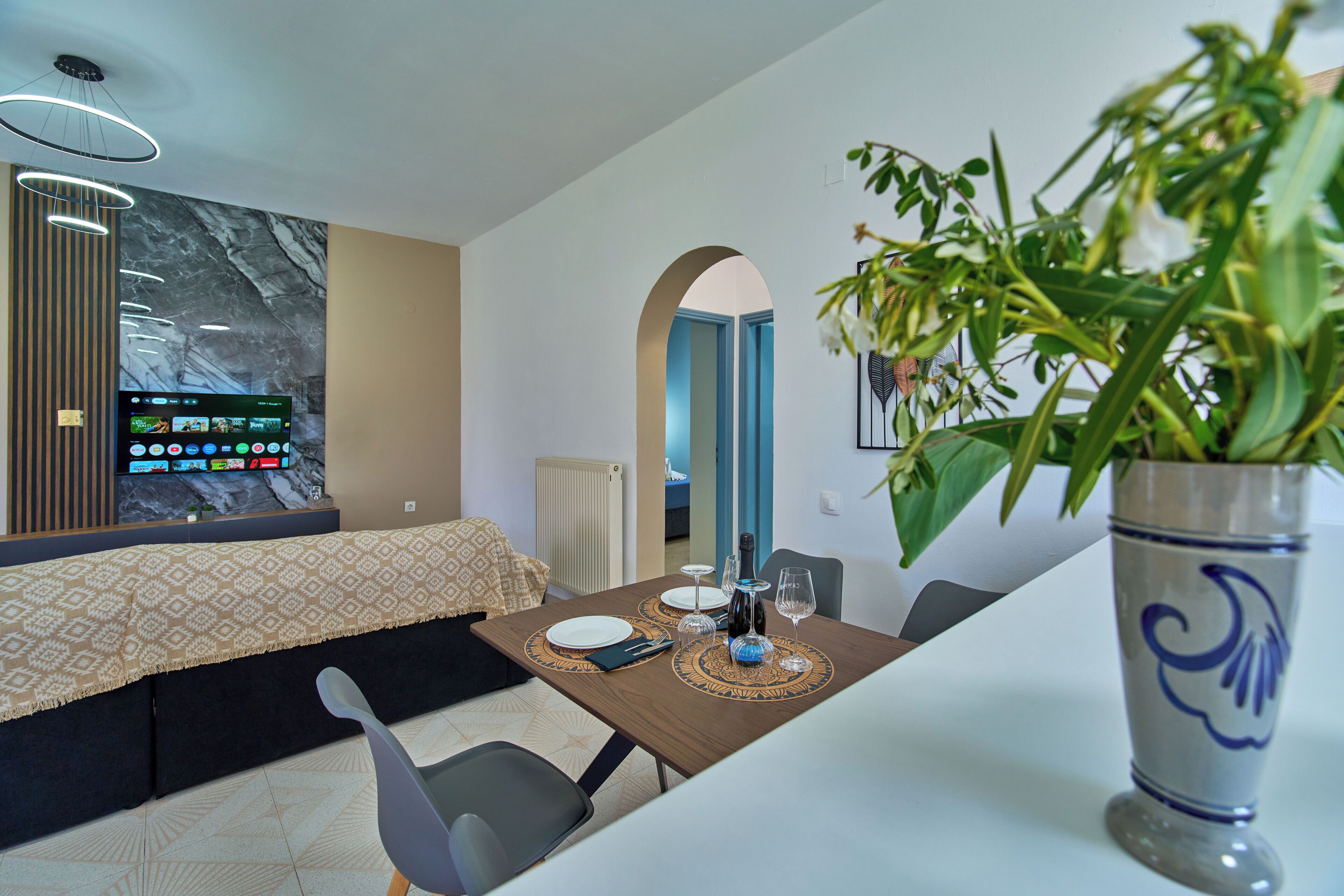 Een moderne eetruimte met een tafel gedekt voor twee personen, een grote vaas met bloemen en een aan de muur bevestigde televisie waarop een menu wordt getoond.