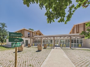 A building with a covered entrance, wooden barrels, and directional signs for 'Bâtiment A' and 'Bar'. - Pavones – Studio for 3 with shared pool (Biscarrosse)