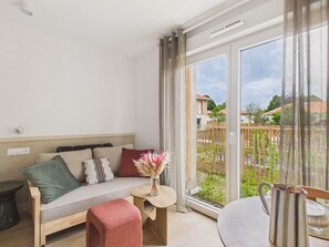 A modern living room with a sofa, a small table with a vase, and a view of a garden through large windows. - Pavones – Studio for 3 with shared pool (Biscarrosse)