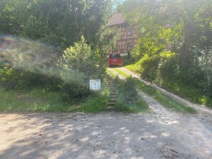 A red car on a dirt path leading to a traditional half-timbered house surrounded by greenery.