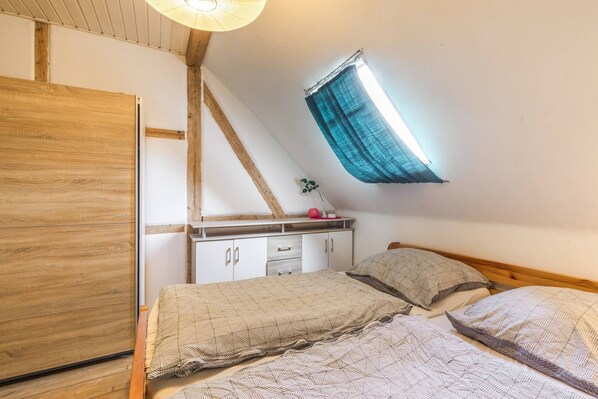 A bedroom with a sloped ceiling, a bed with patterned bedding, a dresser, and a window with a curtain.