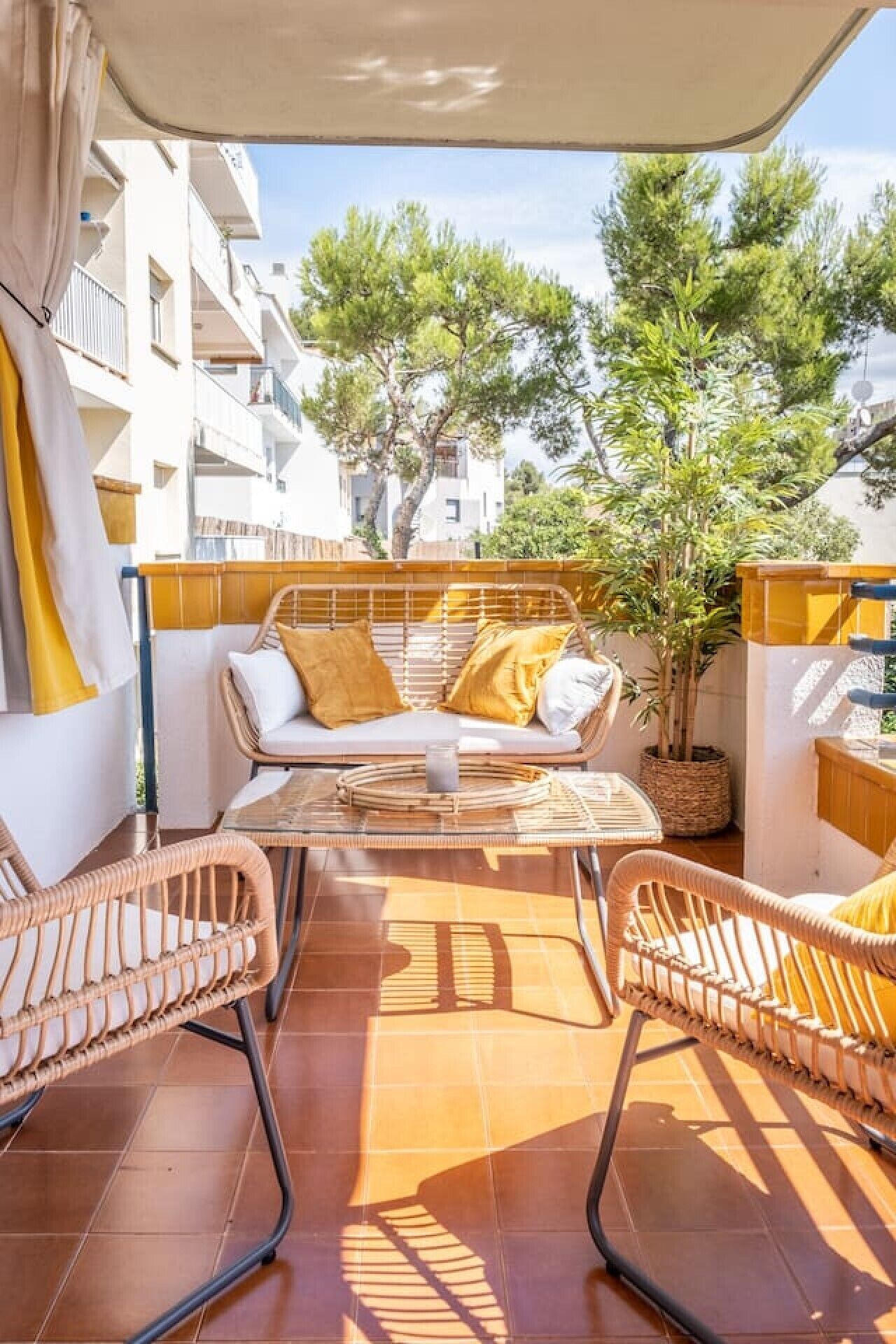 A sunlit outdoor patio with wicker furniture, a glass table, and a view of a residential area with trees and buildings.