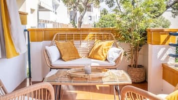 A sunlit outdoor patio with wicker furniture, a glass table, and a view of a residential area with trees and buildings.