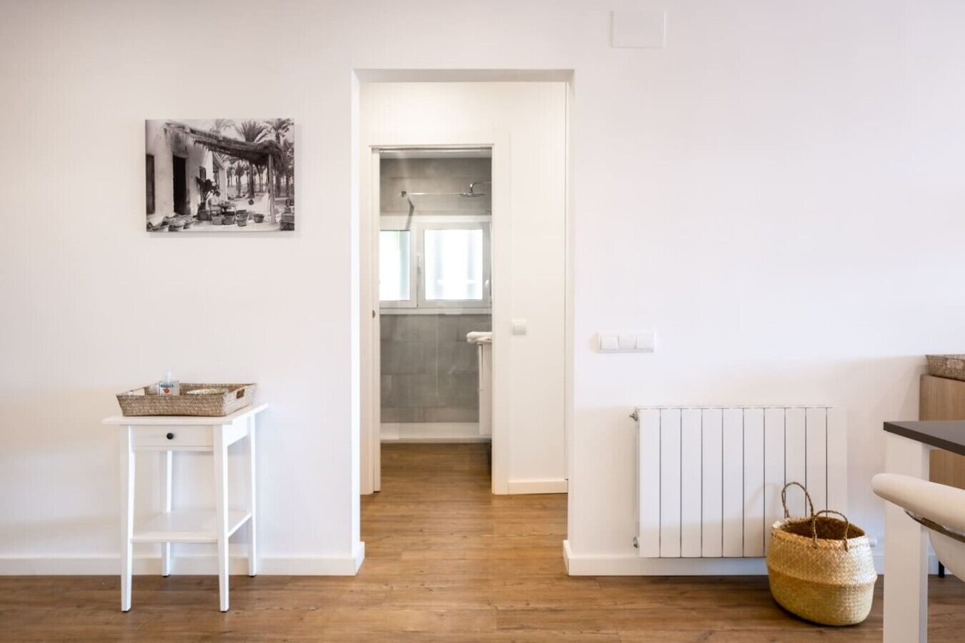 Una habitación con un radiador blanco, una mesita con una cesta, una cesta de mimbre en el suelo y un umbral que da a un baño.