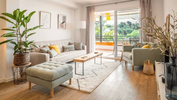 Ein modernes Wohnzimmer mit einer Couch, einem Sessel und einem Hocker, einem Couchtisch und einem großen Fenster mit Blick ins Grüne.