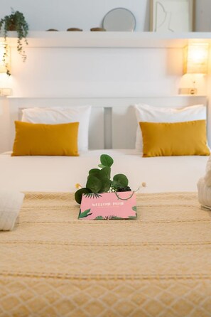 A bed with white and yellow bedding, a pink welcome card, and green plants on a textured rug. - Studio 50m from the Beach | Pedro by Palmera Group (Sitges)