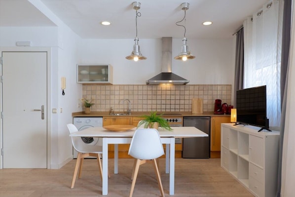 A modern kitchen with white cabinetry, a dining table with chairs, a TV, and pendant lights. - Studio 50m from the Beach | Pedro by Palmera Group (Sitges)
