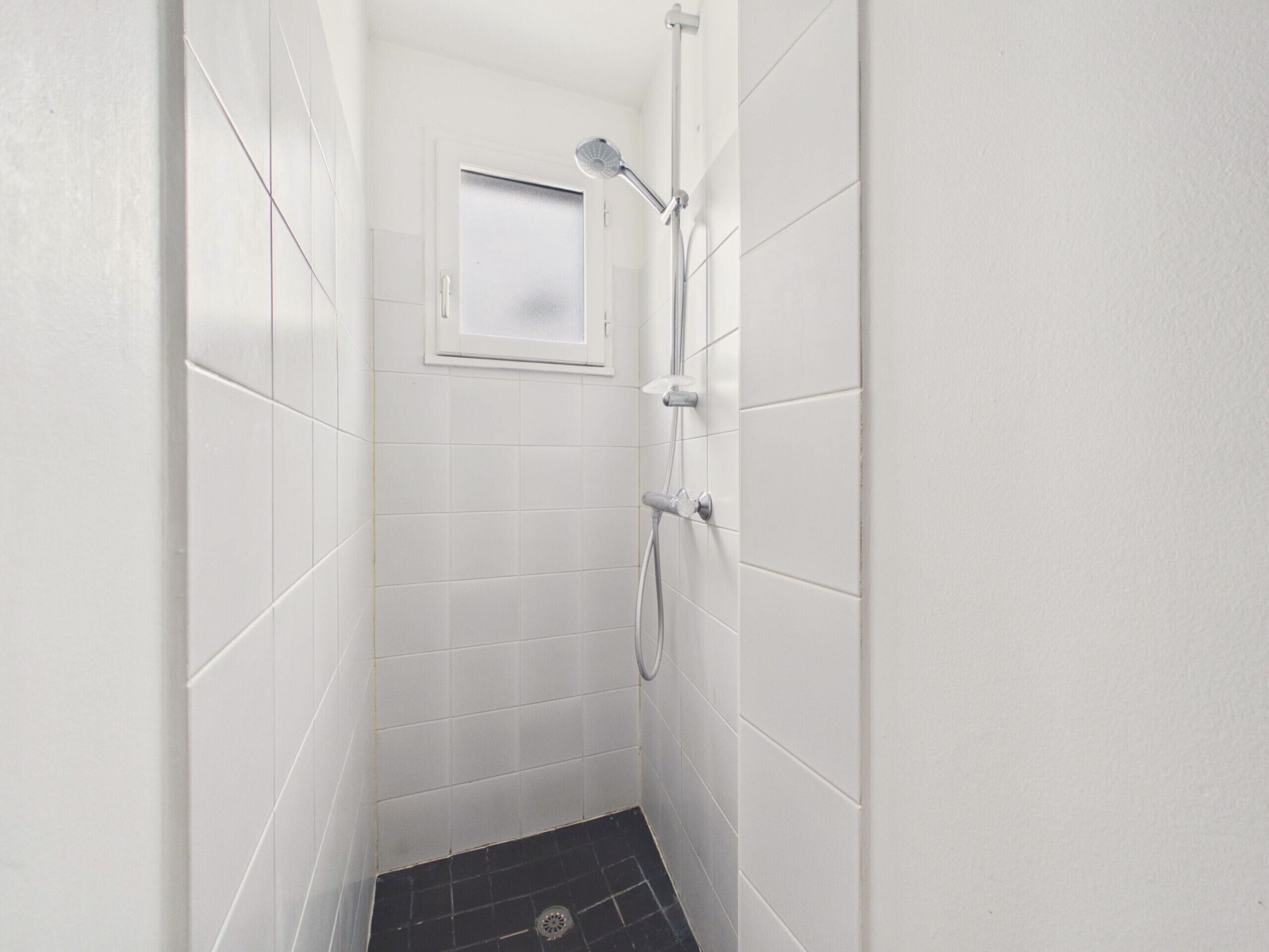 A small, white-tiled shower room with a window, a showerhead, and a handheld shower attachment.