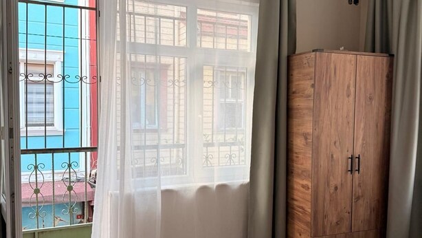 A bedroom with a bed, a wooden wardrobe, a window with curtains, and a view of buildings outside. - walkabout apart (fatih)