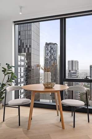 Ein moderner Essbereich mit einem runden Tisch, zwei Stühlen und Blick auf hohe Gebäude durch große Fenster.