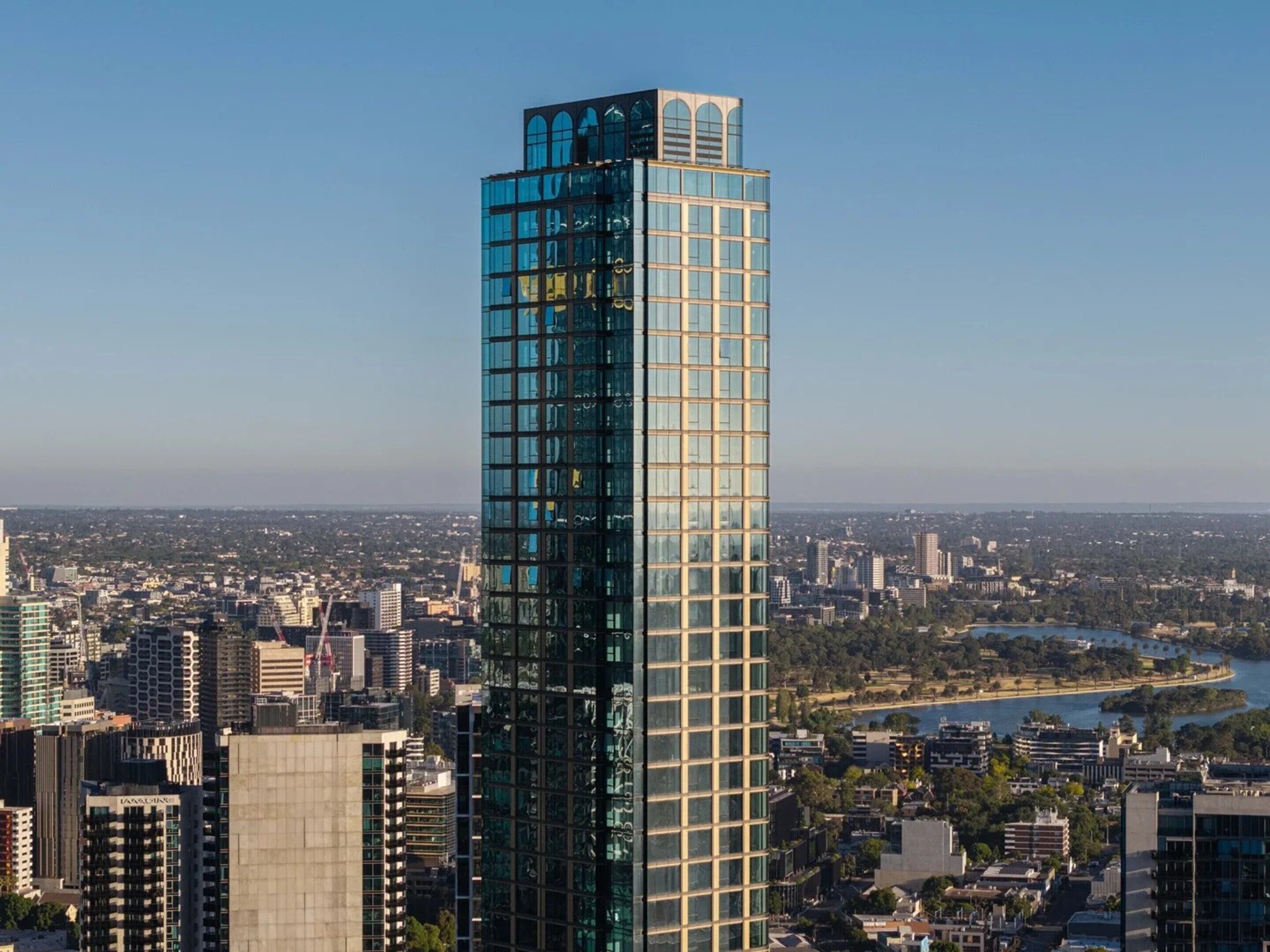 A tall glass skyscraper with a modern design, surrounded by a dense urban area with various buildings and a river in the background.