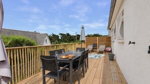 Eine Holzterrasse mit Tisch und Stühlen, ein kleiner Tisch mit Speisen und Getränken und eine weiße Wand mit einem Fenster.
