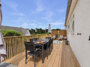 A wooden deck with a table and chairs, a small table with food and drinks, and a white wall with a window. - Near beach, house for 10 with garden (Erquy)