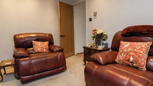 Dos sillones de cuero reclinables con cojines estampados con motivos florales, una pequeña mesita de madera con un florero y una puerta de madera en una habitación de colores neutros.