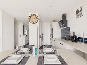 A modern kitchen with a large wall clock, a dining table set for four, and a coffee machine. - Sea-view balcony & beach access - Sleeps 4 (Saint-Hilaire-de-Riez)