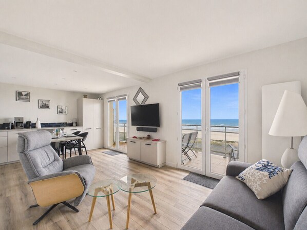 A modern living room with a sofa, a glass coffee table, and a TV. There is a balcony with a view of the ocean. - Sea-view balcony & beach access - Sleeps 4 (Saint-Hilaire-de-Riez)