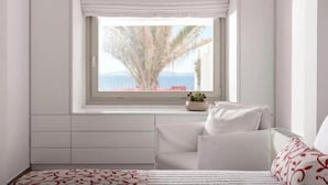 A bedroom with a large window, a bed, a bedside table, and a view of palm trees and the sea. - O Lofos DELOS Villa with Sea View /Private Pool (Mykonos)