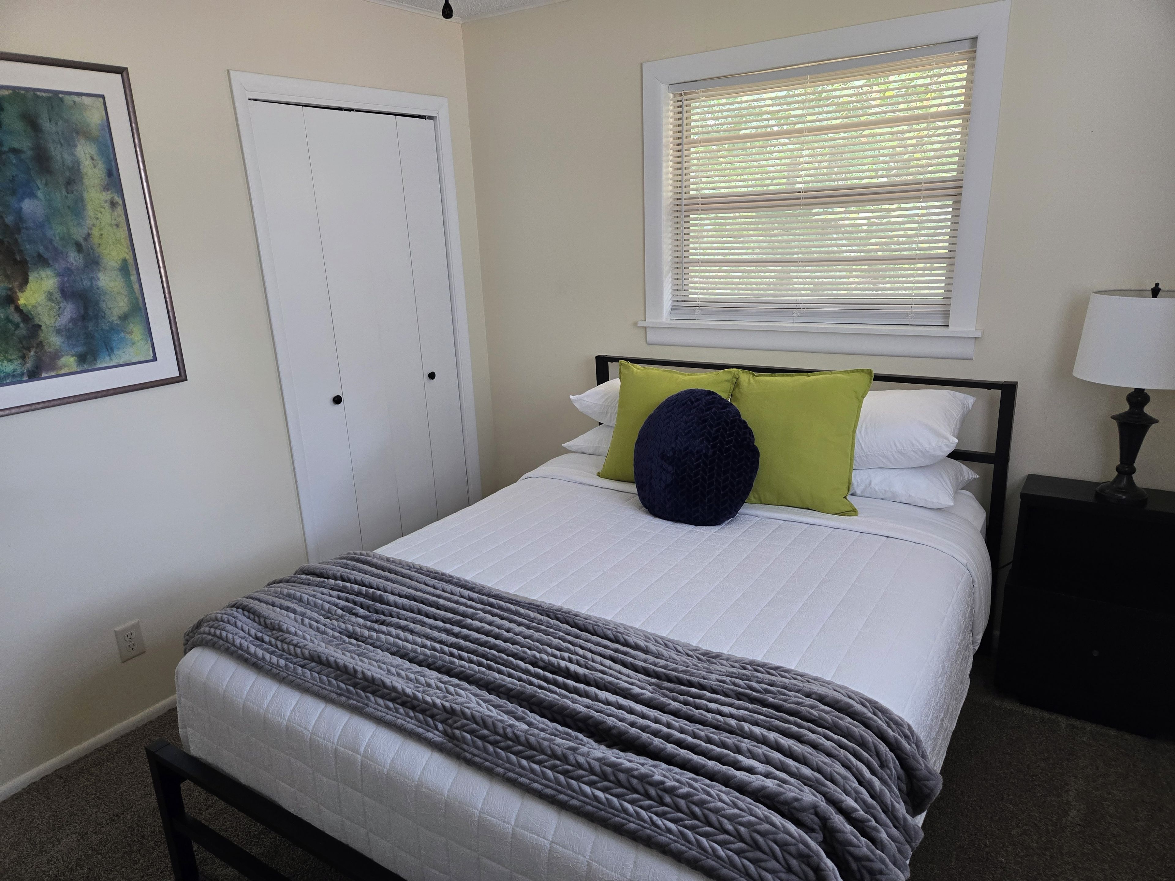 Une chambre à coucher comprenant un lit, une table de chevet, une lampe, une fenêtre avec des stores, une porte et un tableau encadré accroché au mur.