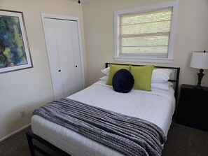 Ein Schlafzimmer mit einem Bett, einem Nachttisch, einer Lampe, einem Fenster mit Jalousien, einer Tür und einem gerahmten Bild an der Wand.