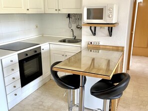 A compact kitchen with a microwave, oven, sink, and two bar stools. - Beachfront Apartment with Pool, Terrace & Wi-Fi – Ideal for Families (La Pineda)