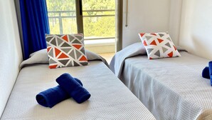 Zwei Betten mit gemusterten Kissen und aufgerollten Handtüchern, ein Fenster mit blauen Vorhängen und Blick ins Grüne.