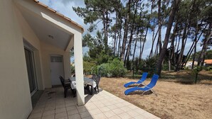 Un patio cubierto con mesa y sillas, un área vallada con sillones de descanso, y vistas a árboles y un edificio distante.