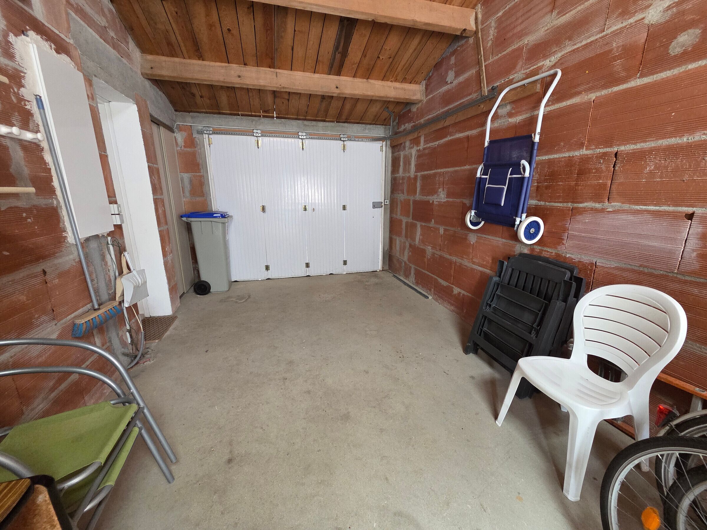 A room with a concrete floor, a wooden ceiling, a white door, a blue chair, a white plastic chair, and a bicycle.