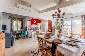 A spacious living area with a dining table set for four, a kitchen in the background, and a large window providing natural light. - Villa 'Micoco' with Private Terrace, Wi-Fi and Air Conditioning (Gujan-Mestras)