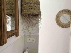 Ein Badezimmer mit einem spiegelnden Holzboden, einer gewebten Wanddekoration, einem runden Spiegel mit Fransen und einem Zahnputzhalter mit einer Zahnbürste.