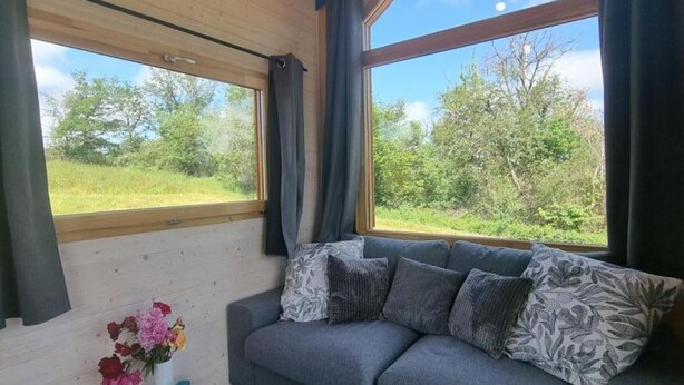 Ett mysigt rum med en grå soffa, ett litet träbord med en vas med blommor och stora fönster med utsikt över grönskan.