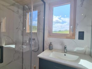 A bathroom with a glass shower enclosure, a sink with a mirror above it, and a view of the sky through a window.