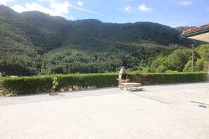 Ein gepflasterter Außenbereich mit Grill, Bänken und Blick auf einen bewaldeten Berg.